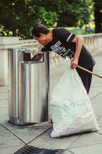 A man and two children are engaged in a beach cleanup during daytime, with the man wearing a red t-shirt, light blue shorts, and a black cap, holding a white garbage bag in one hand. The young boy, dr