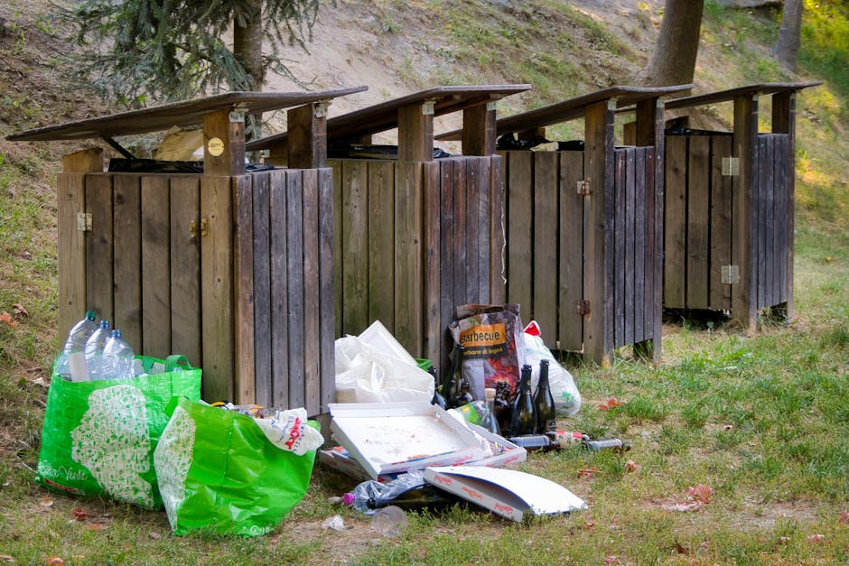 A large green waste disposal bin filled with a mixture of cardboard boxes, including some flattened and others partially intact, alongside pieces of packaging and paper. The cardboard varies in colour